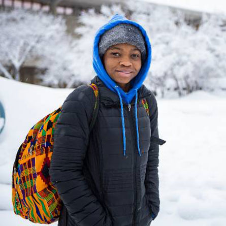 A student in winter gear smiles in front of a snowy plaza on the UAF campus